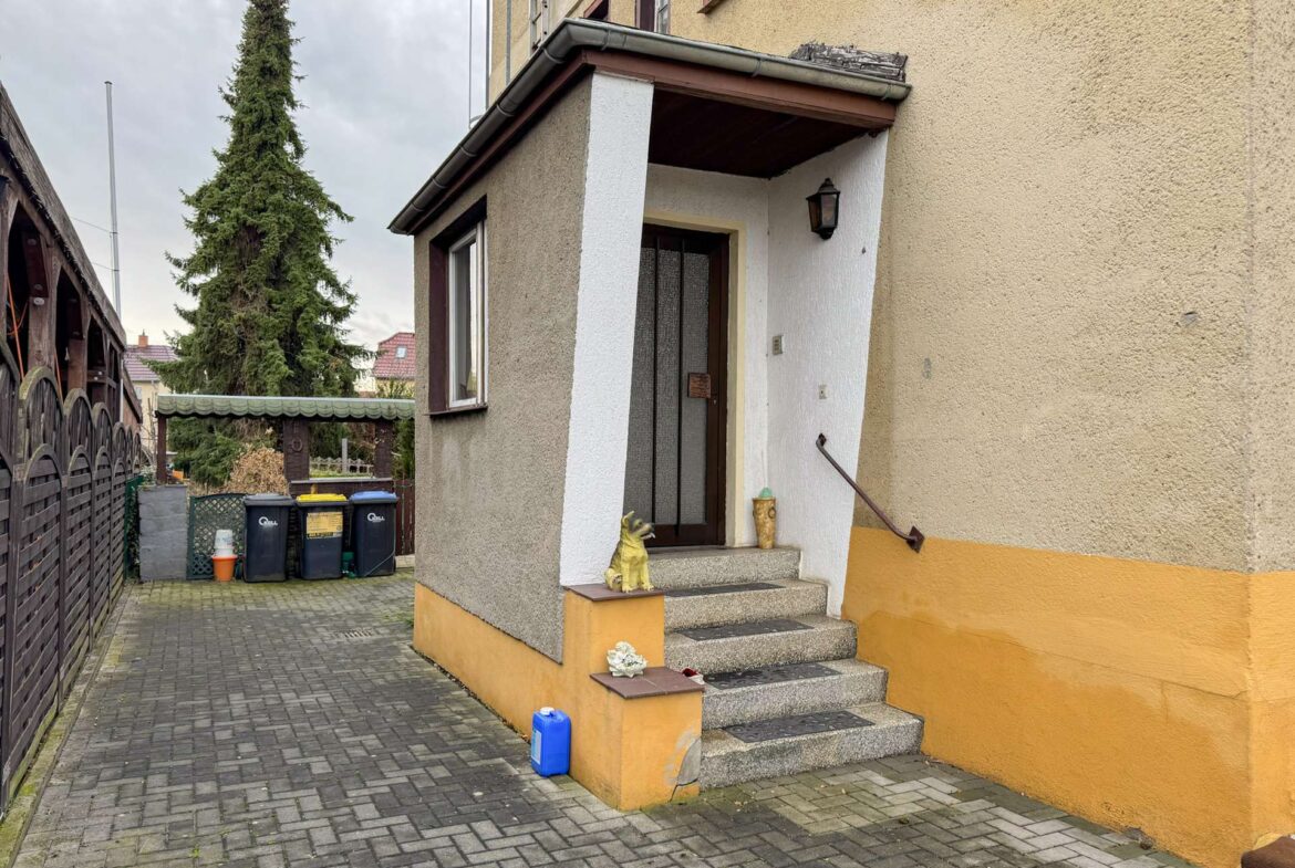 Front entrance of a beige house with an orange base; concrete steps, barred door, a small dog statue on the step, and a lantern on the wall.