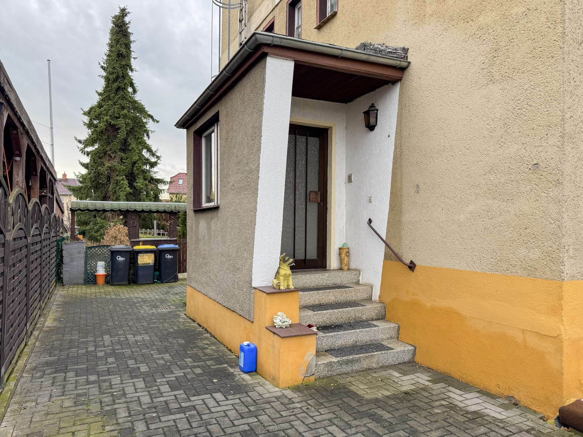 Front entrance of a beige house with an orange base; concrete steps, barred door, a small dog statue on the step, and a lantern on the wall.