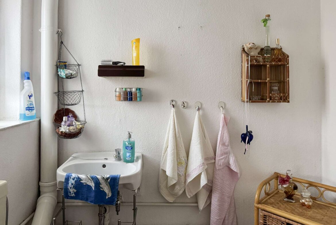 Bathroom corner with a white sink, pink and white towels on hooks, and open storage racks holding toiletries and cleaning products on the left and right.