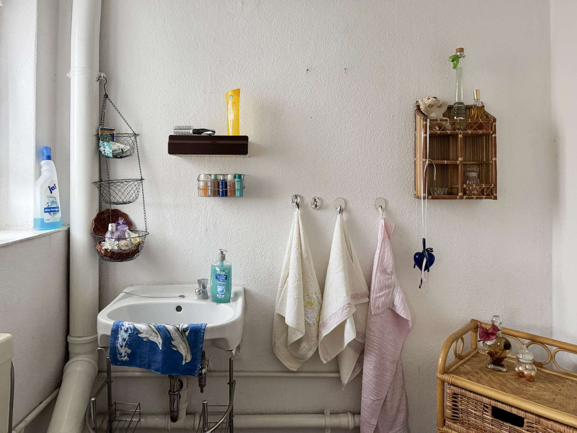 Bathroom corner with a white sink, pink and white towels on hooks, and open storage racks holding toiletries and cleaning products on the left and right.