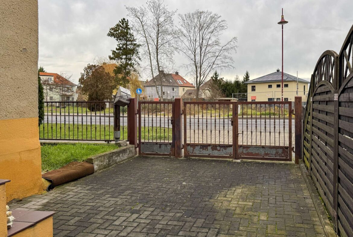 Gate blocks a cobblestone courtyard with a yellow-orange wall on the left and a wooden fence on the right, against a backdrop of houses and leafless trees under a cloudy sky.