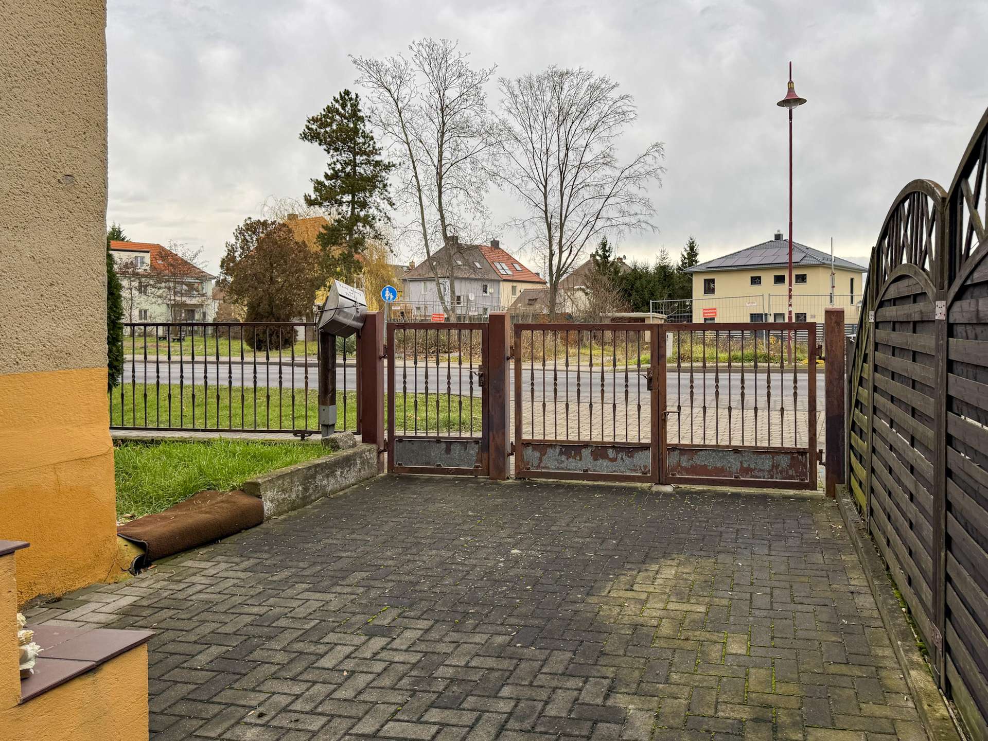 Gate blocks a cobblestone courtyard with a yellow-orange wall on the left and a wooden fence on the right, against a backdrop of houses and leafless trees under a cloudy sky.