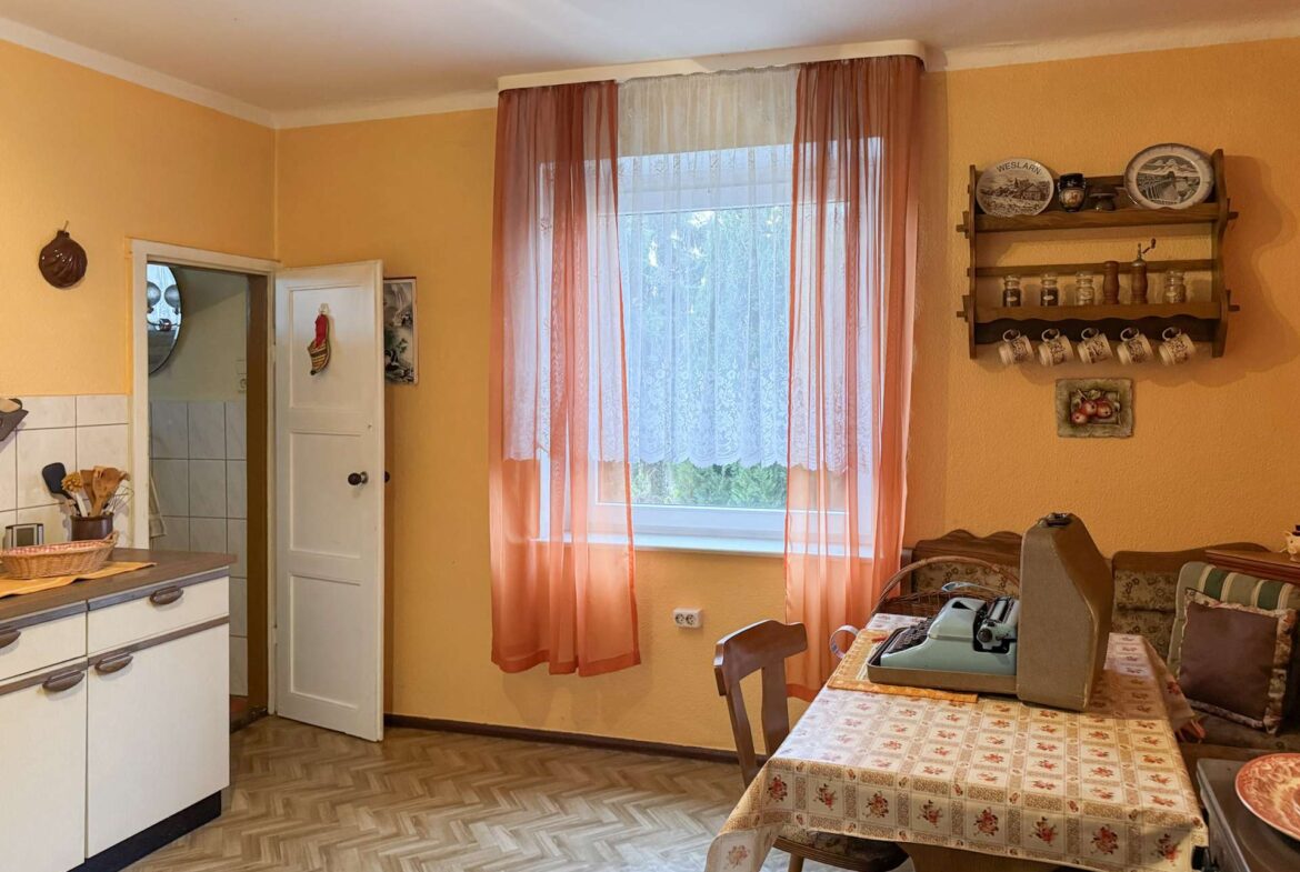 Kitchen/dining area with peach walls, pink sheer curtains, and a window letting in light; a wooden shelf with plates and mugs on the right.