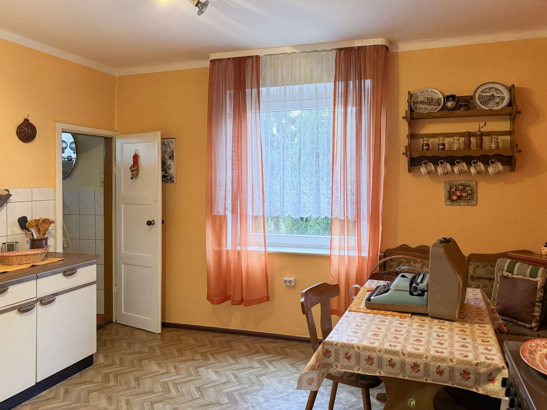 Kitchen/dining area with peach walls, pink sheer curtains, and a window letting in light; a wooden shelf with plates and mugs on the right.