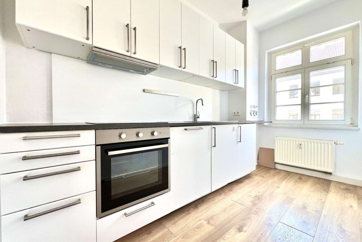 White modern kitchen with tall white cabinets, built-in oven, metallic handles, and a large window letting in daylight by a radiator.