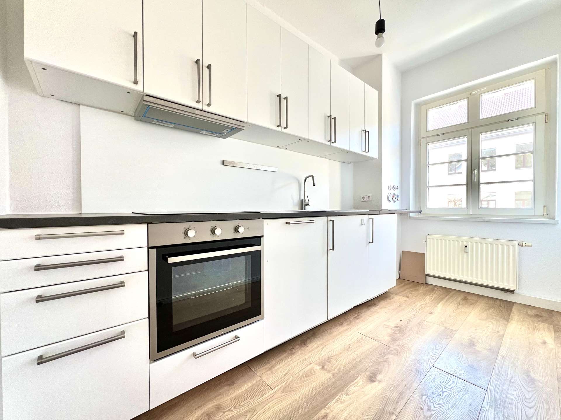 White modern kitchen with tall white cabinets, built-in oven, metallic handles, and a large window letting in daylight by a radiator.