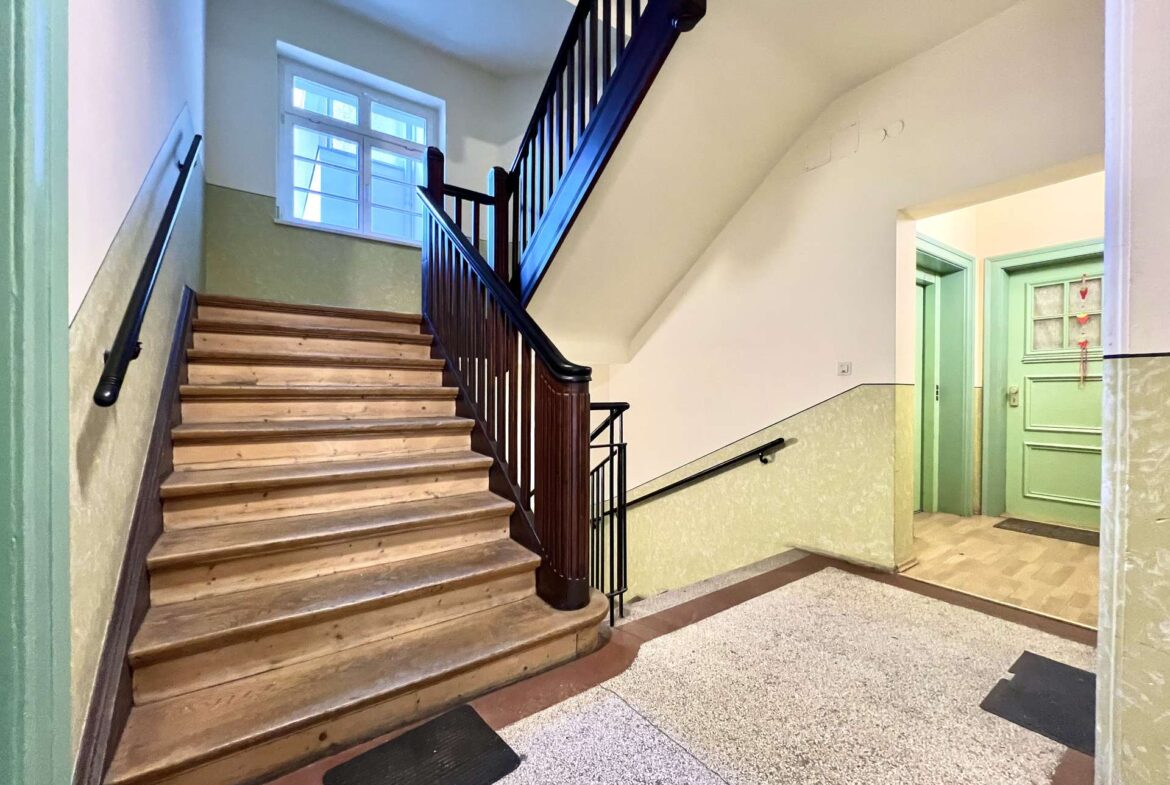 Interior stairwell with wooden staircase and black handrail, green doors to the right, light cream walls, and speckled tiled floor.