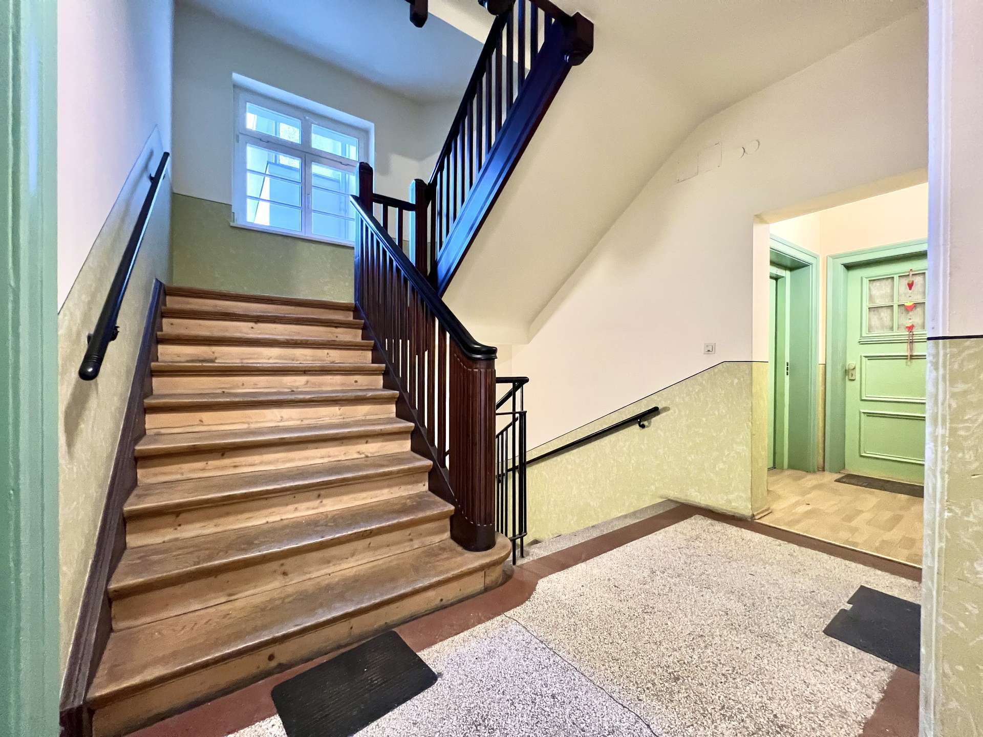 Interior stairwell with wooden staircase and black handrail, green doors to the right, light cream walls, and speckled tiled floor.