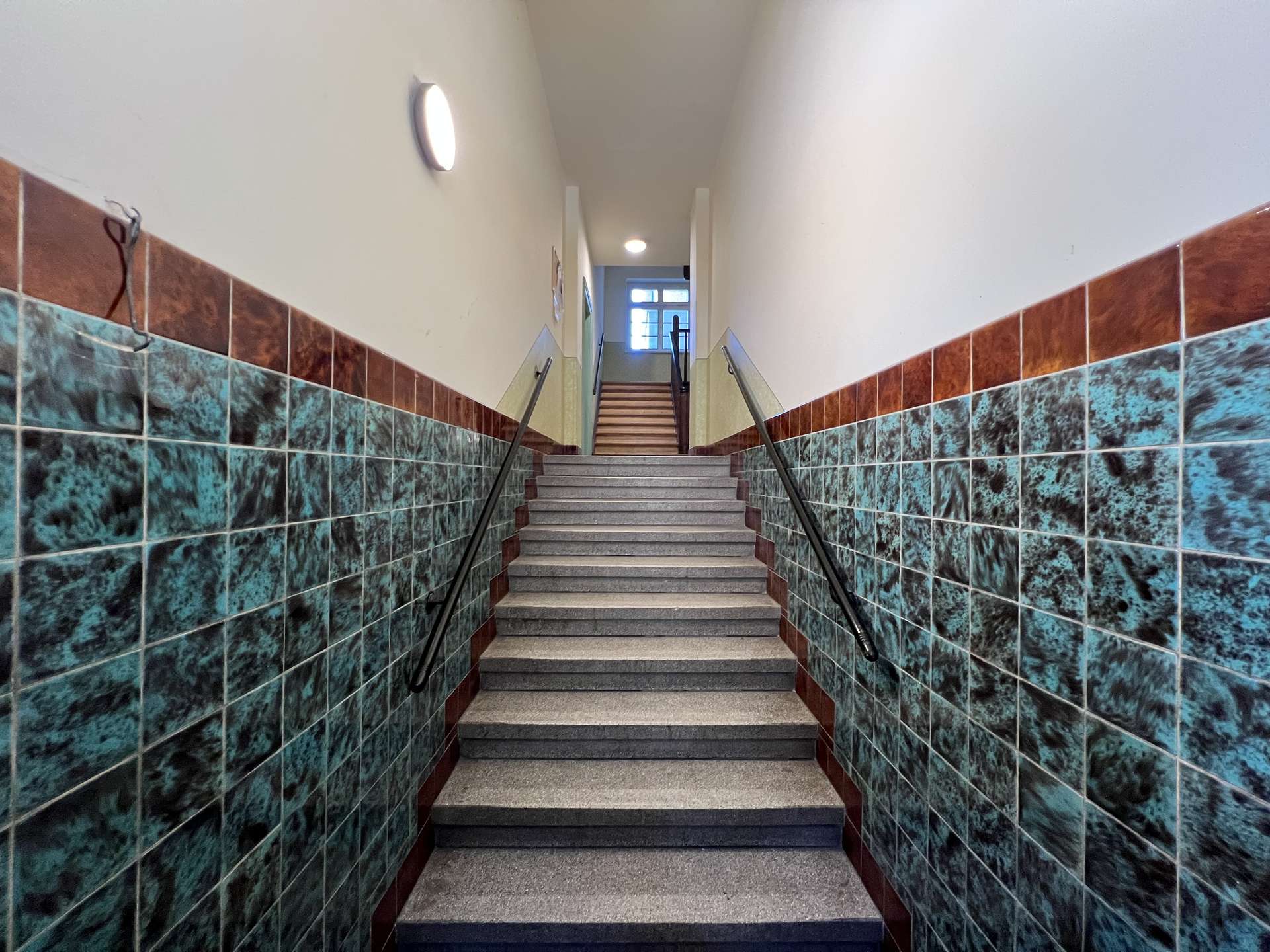 Indoor stairwell with teal marbled wall tiles, brown trim, and metal handrails leading to a window at the top.