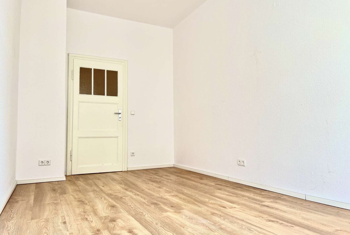 Empty white room with light wood floor and a white door on the left wall.