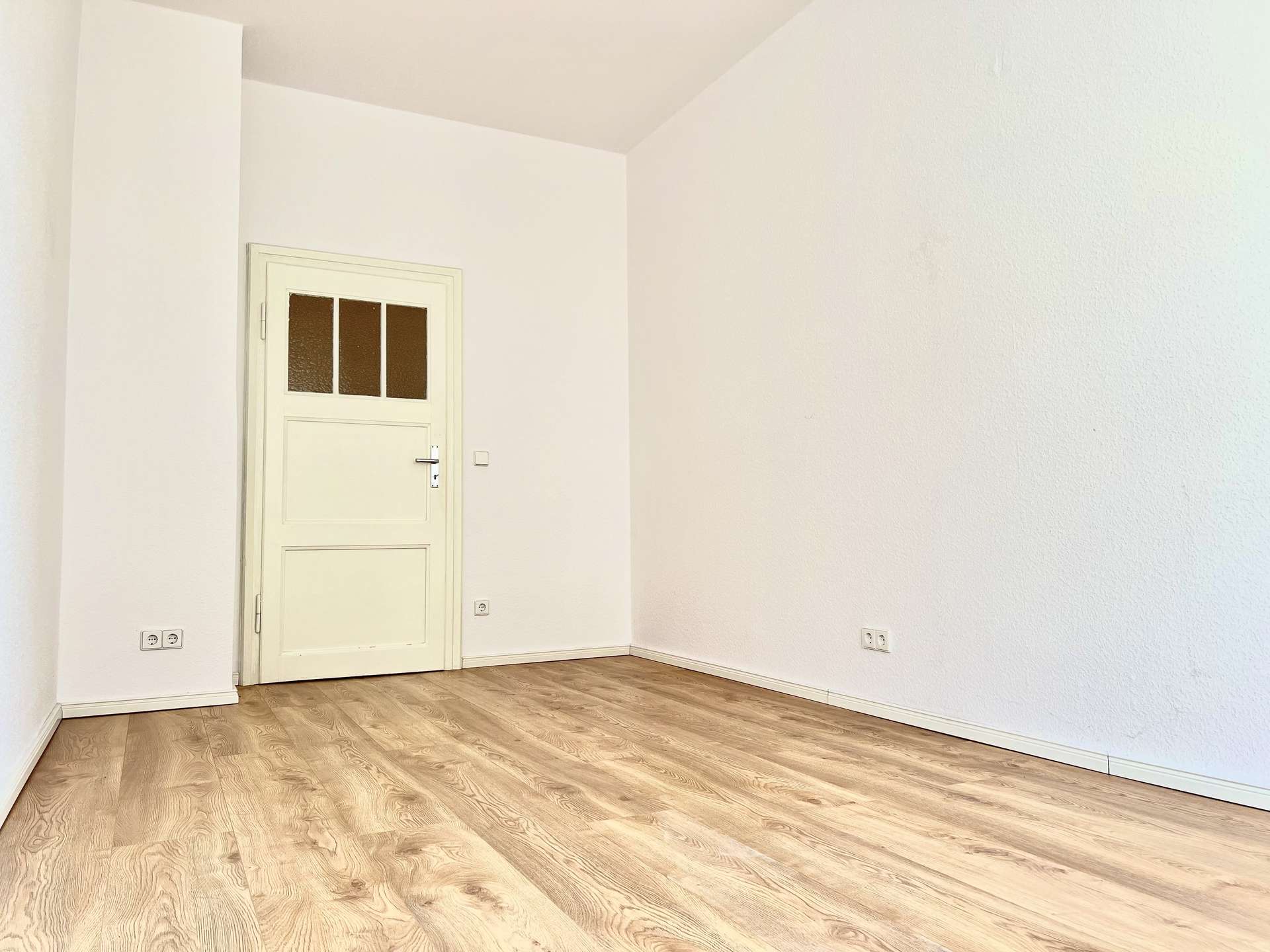 Empty white room with light wood floor and a white door on the left wall.