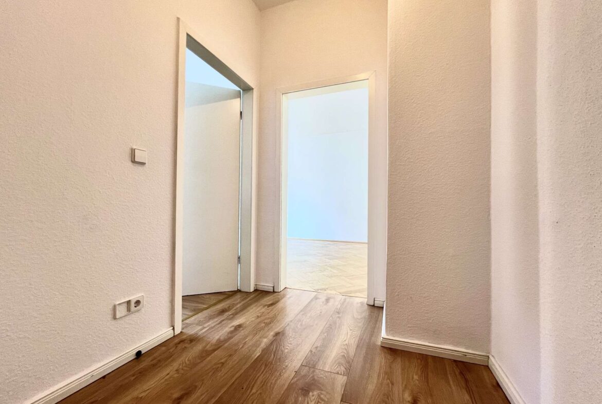 White-walled hallway with wood floors and an open doorway to a bright room at the end.