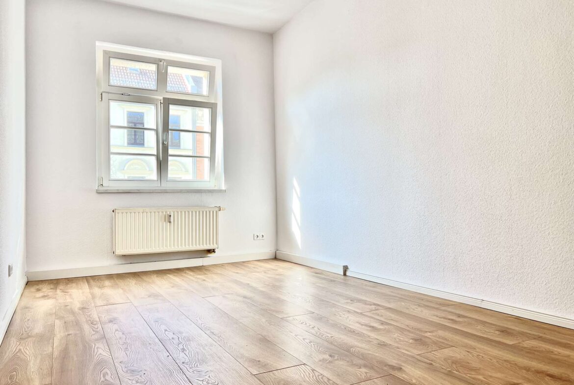Empty bright room with a large window, radiator beneath it, and light wood floor panels.