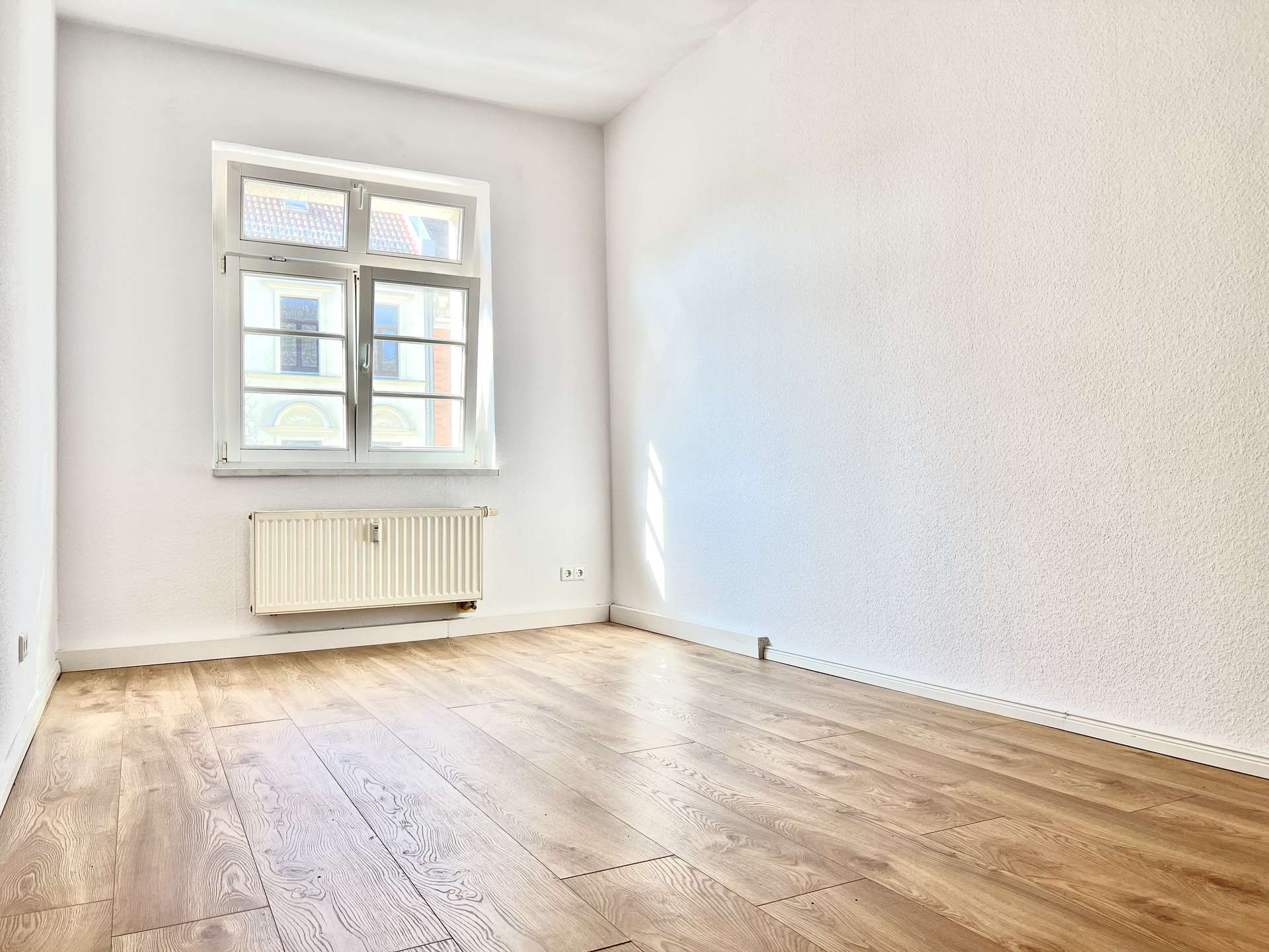 Empty bright room with a large window, radiator beneath it, and light wood floor panels.