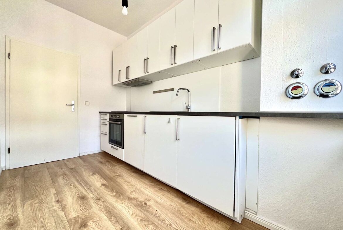 White modern kitchen with tall white cabinets, built-in oven, and a stainless steel sink along a black countertop; bare lightbulb hangs from the ceiling.
