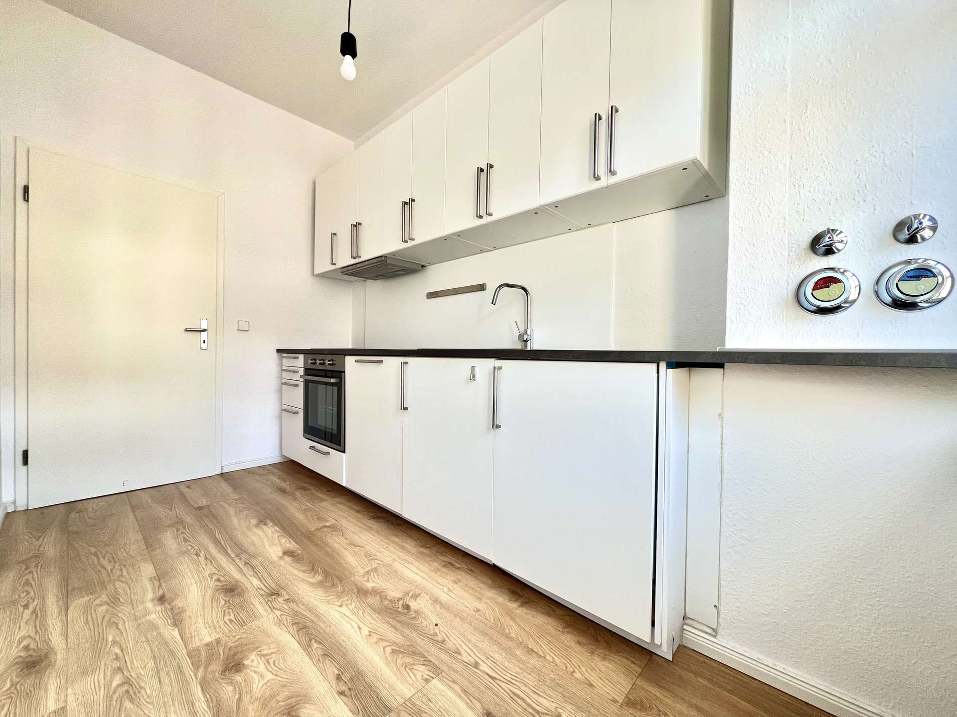 White modern kitchen with tall white cabinets, built-in oven, and a stainless steel sink along a black countertop; bare lightbulb hangs from the ceiling.