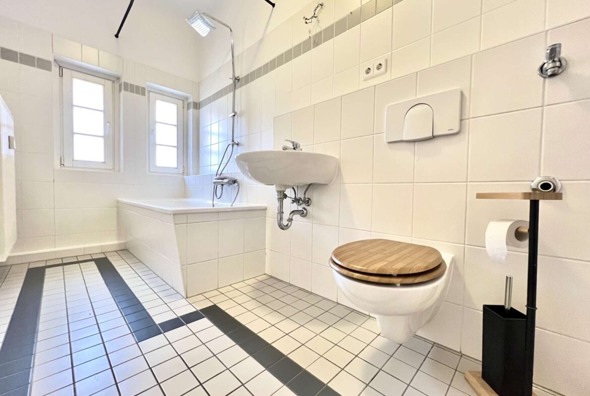 Bright white bathroom with bathtub and rain shower, wall-mounted toilet with wooden seat, and a sink against tiled walls.
