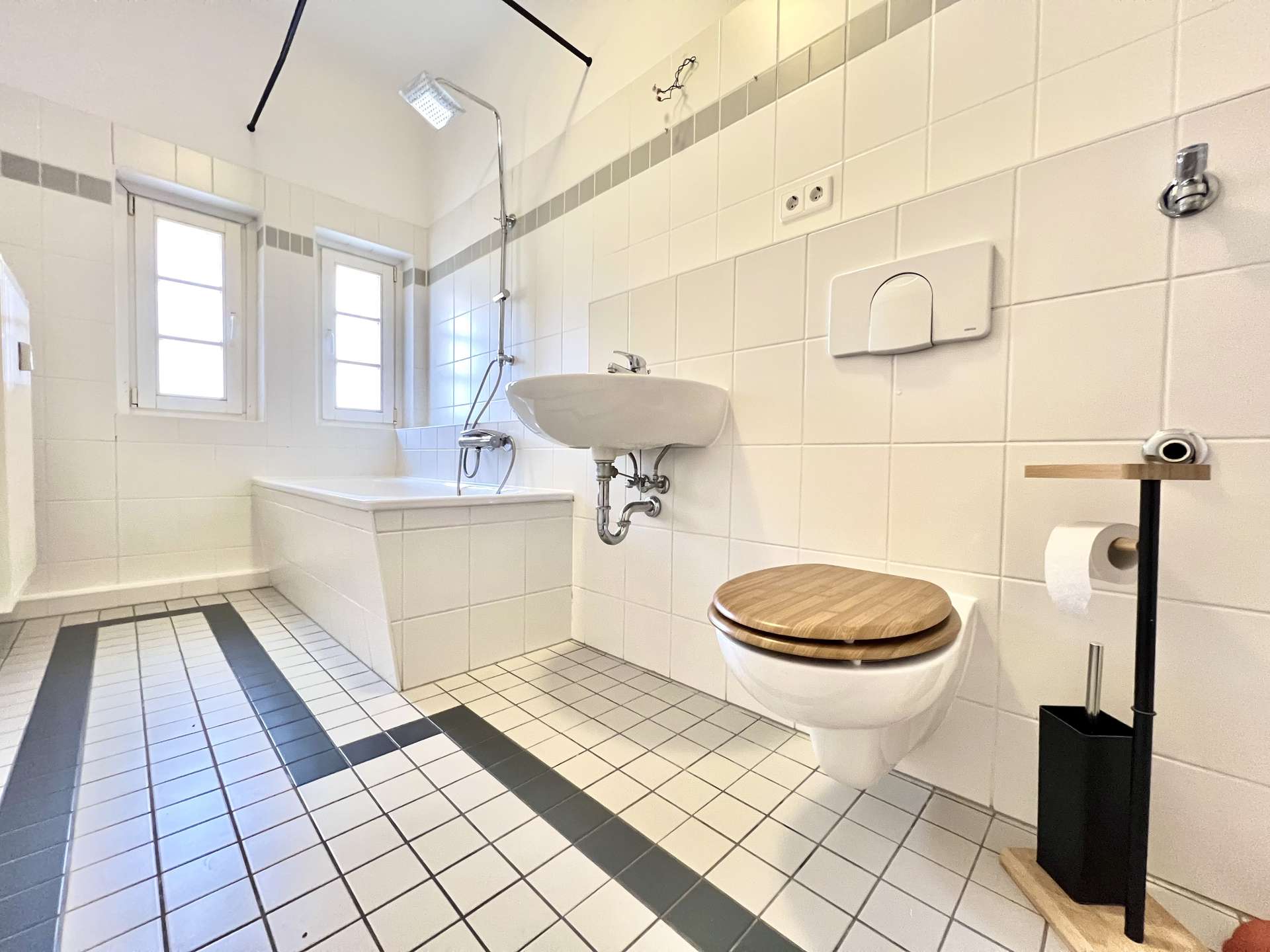 Bright white bathroom with bathtub and rain shower, wall-mounted toilet with wooden seat, and a sink against tiled walls.