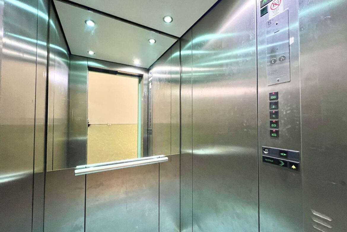 Interior of a stainless steel elevator cab with bright ceiling lights and mirrored walls, door frame visible ahead.