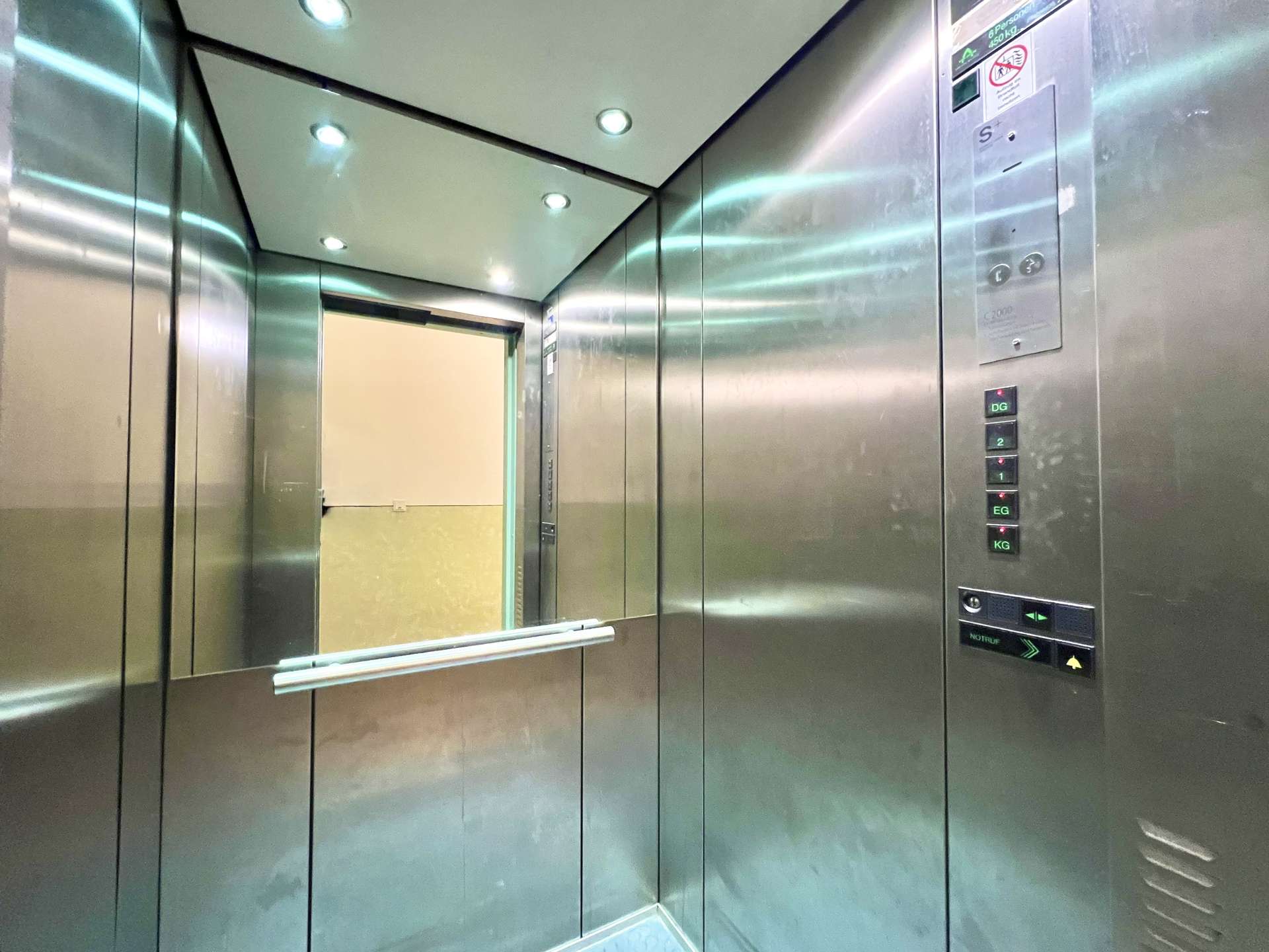 Interior of a stainless steel elevator cab with bright ceiling lights and mirrored walls, door frame visible ahead.