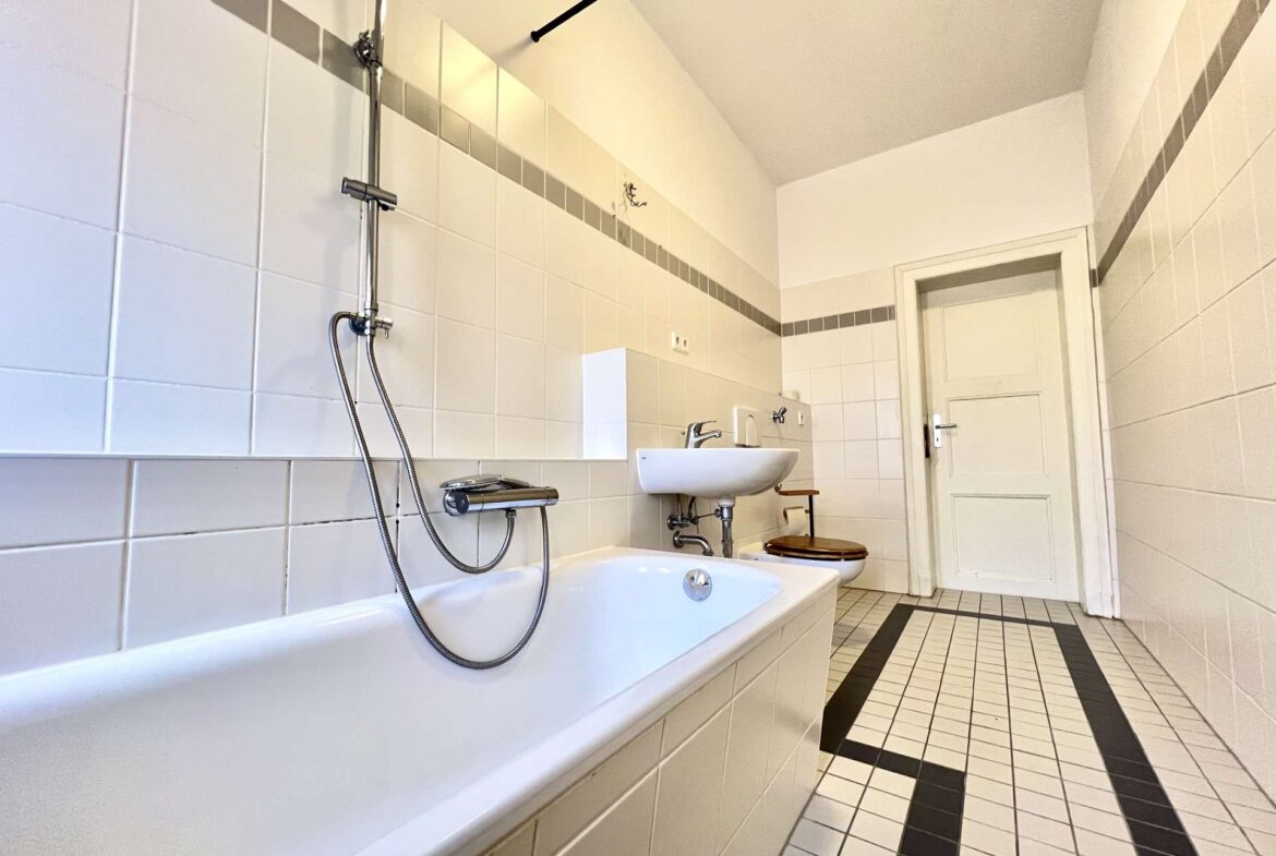 Bright bathroom with white wall tiles, a built-in bathtub and handheld shower, plus a wall-mounted sink and toilet along a tiled wall.