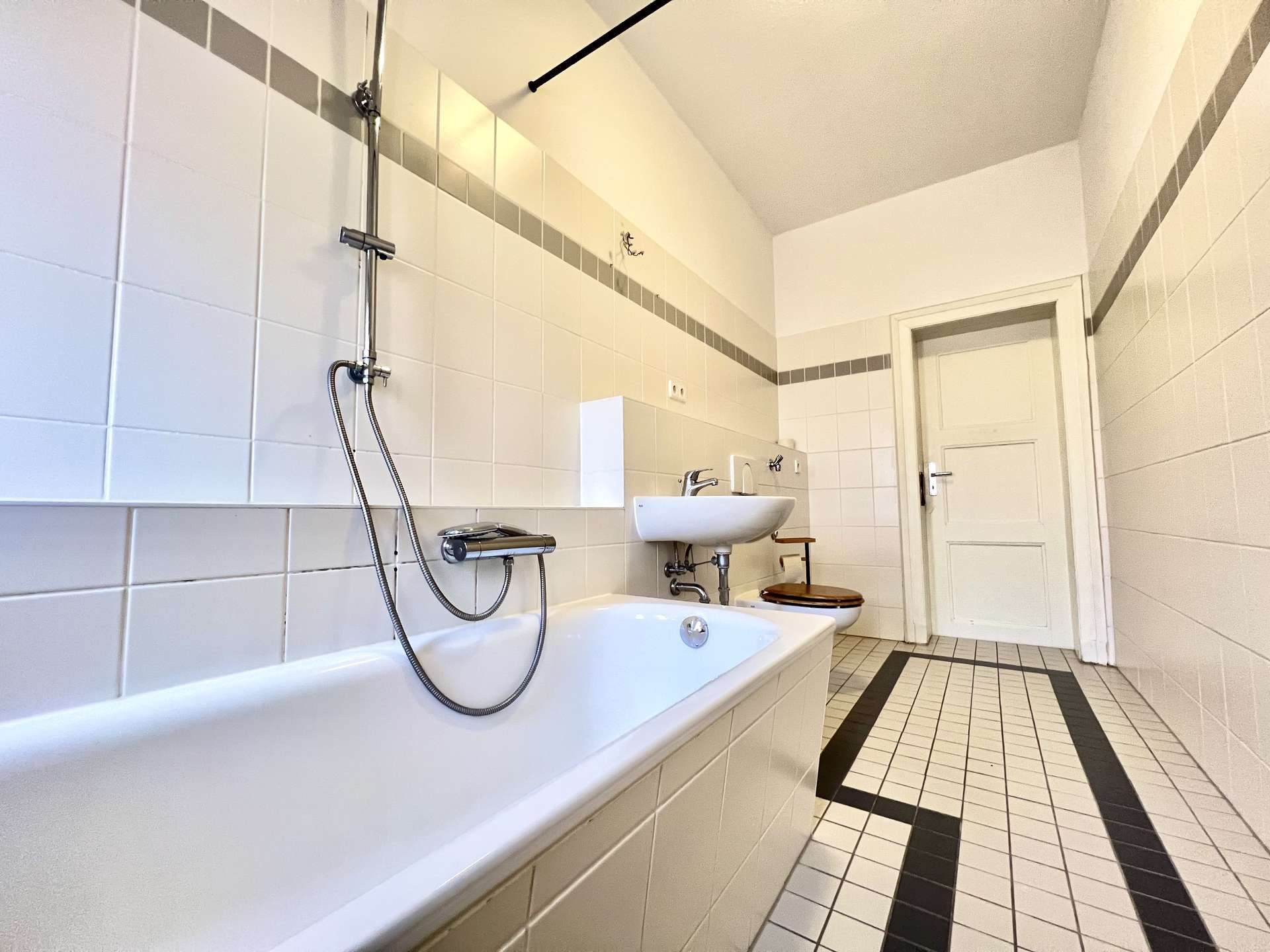 Bright bathroom with white wall tiles, a built-in bathtub and handheld shower, plus a wall-mounted sink and toilet along a tiled wall.