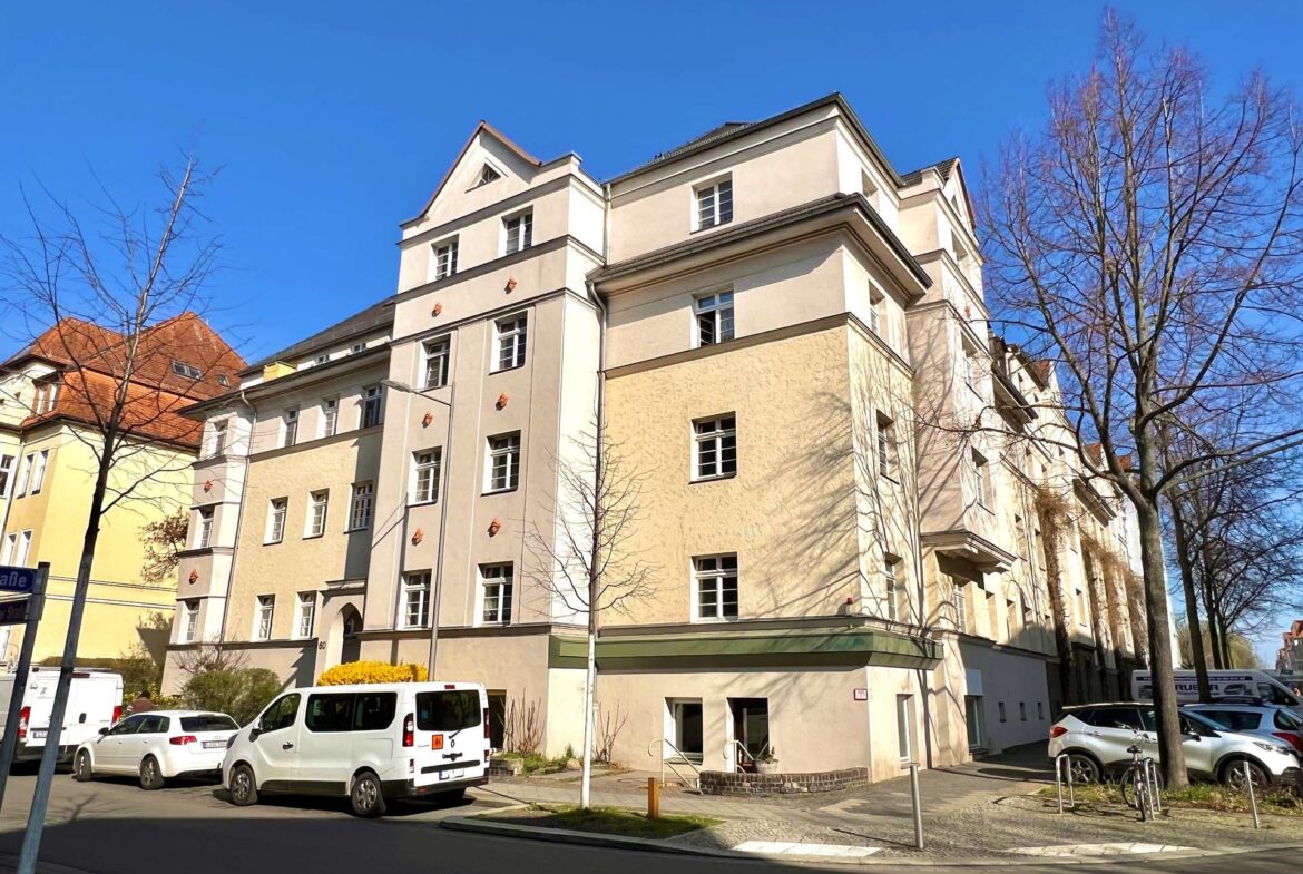 Street view of a pale beige multi-story apartment building with bare trees and parked cars against a clear blue sky.