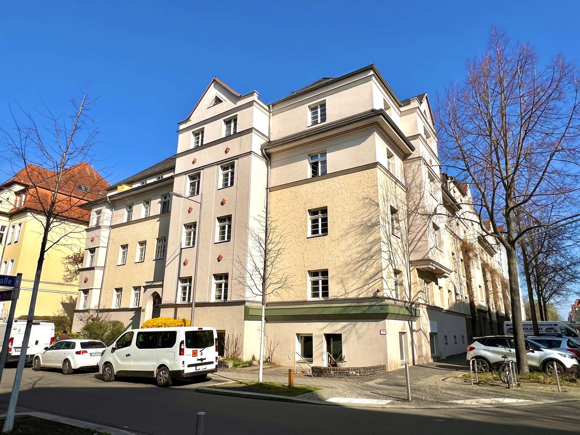 Street view of a pale beige multi-story apartment building with bare trees and parked cars against a clear blue sky.