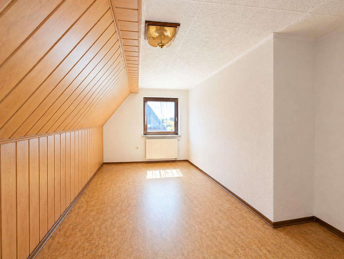 Empty attic room with slanted wooden paneling on the left wall and ceiling, white side wall, small window with radiator beneath it, and cork flooring.