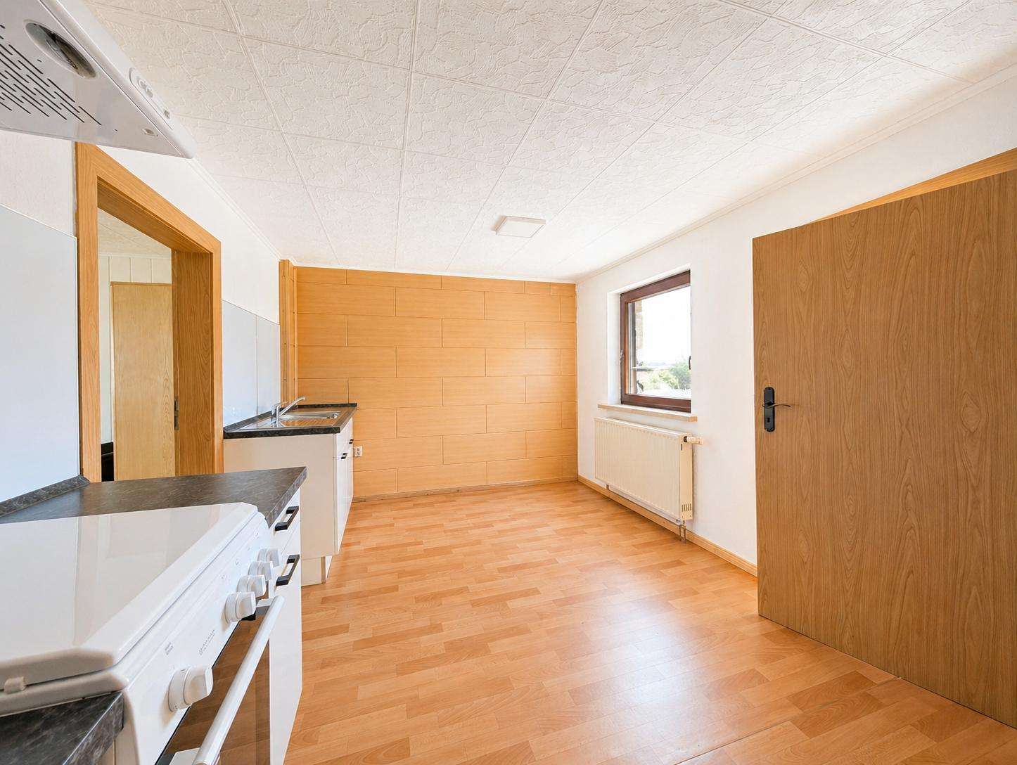 Bright empty kitchen with white stove, dark countertops, wooden door, and a large window.
