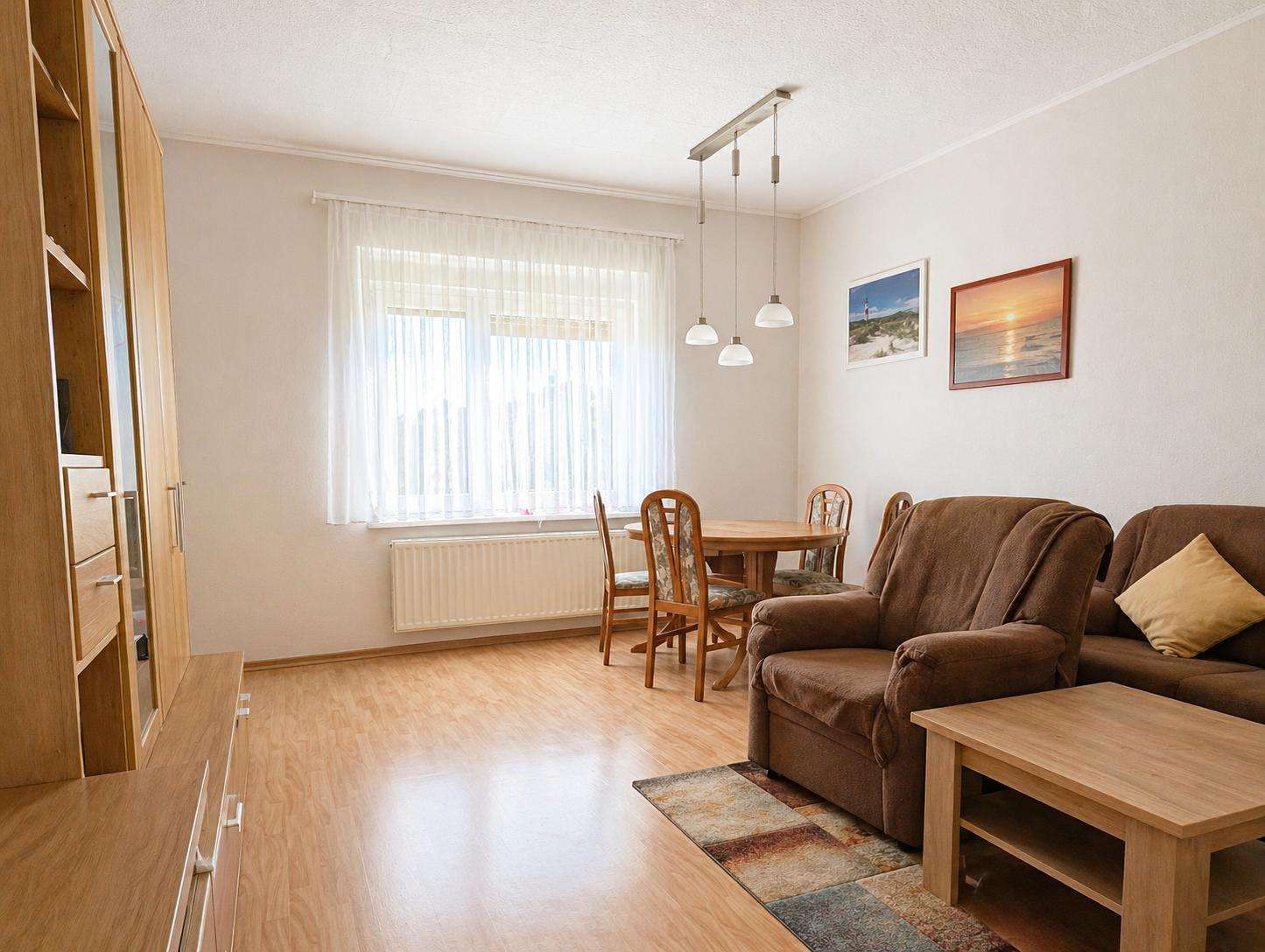 Living room with a brown sofa set, wooden coffee table, and a round dining table near a large window with sheer white curtains.