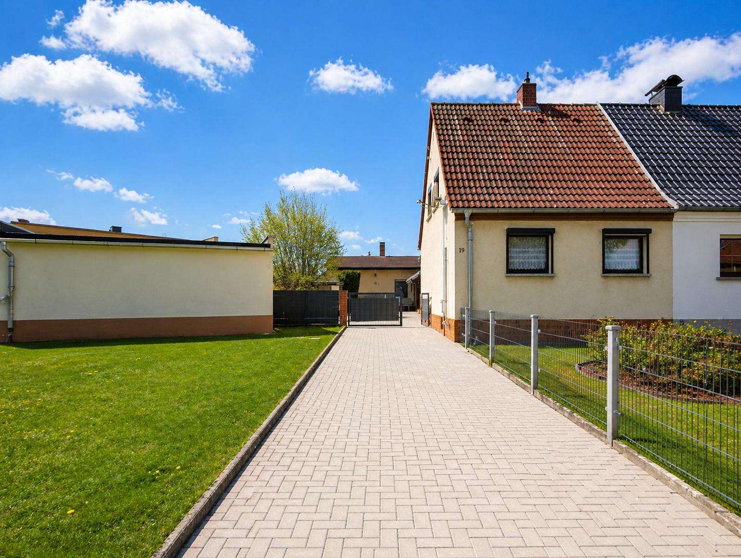 Paved driveway leading to a beige house with a red-tiled roof, black gate, and green lawn on the left under a blue sky.