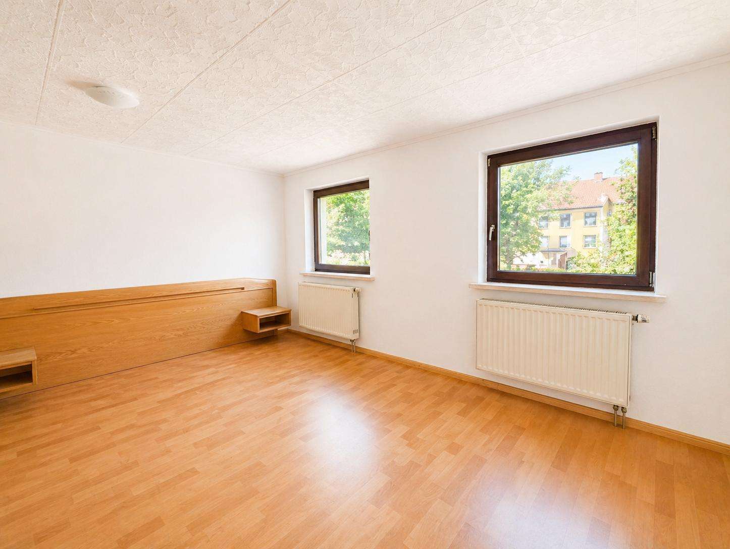 Empty sunlit room with light wooden floor, a built-in wooden platform bed along the left wall, and two windows with radiators beneath them.