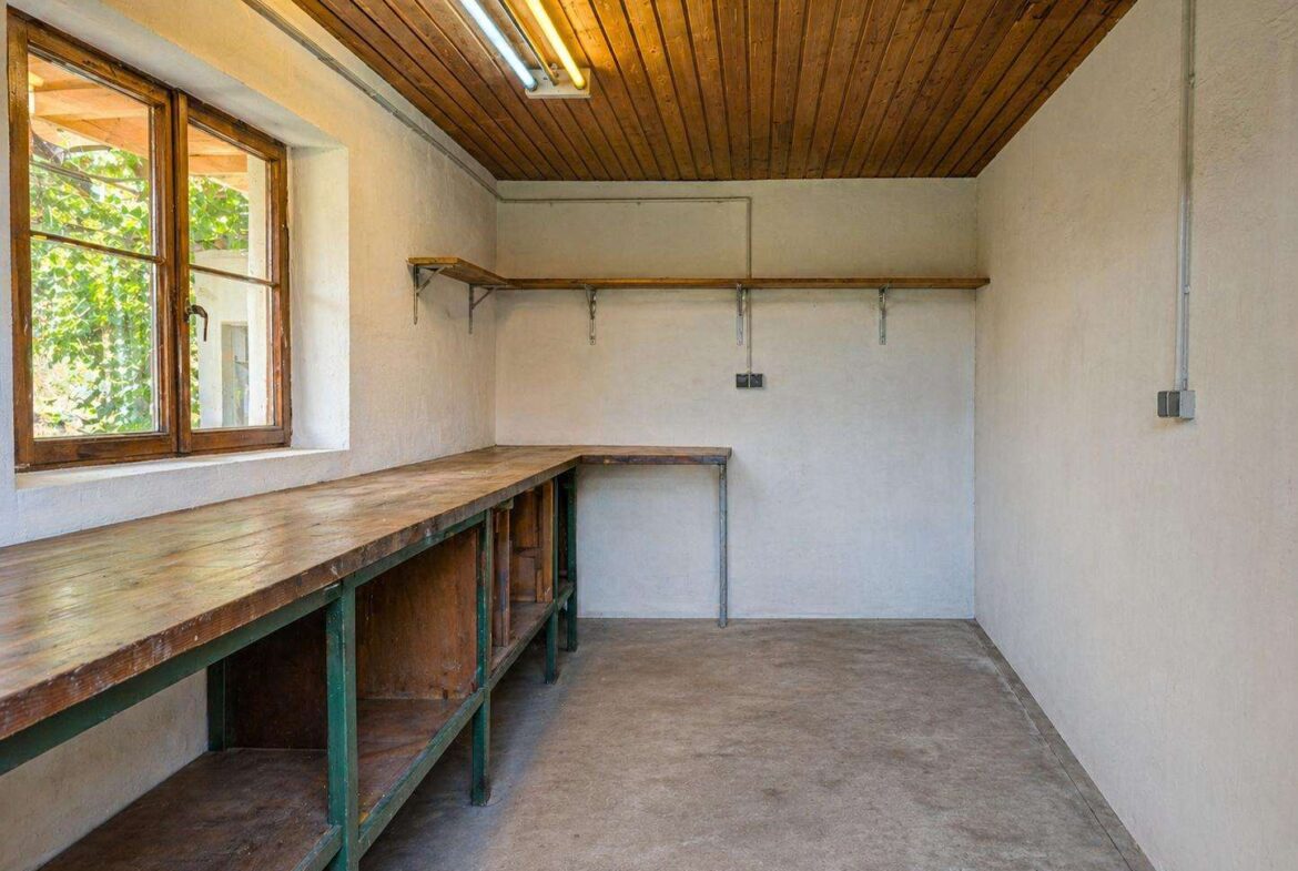 Long wooden workbench runs along the left wall in a simple workshop, with shelves underneath, a window, and an empty, light-filled room.