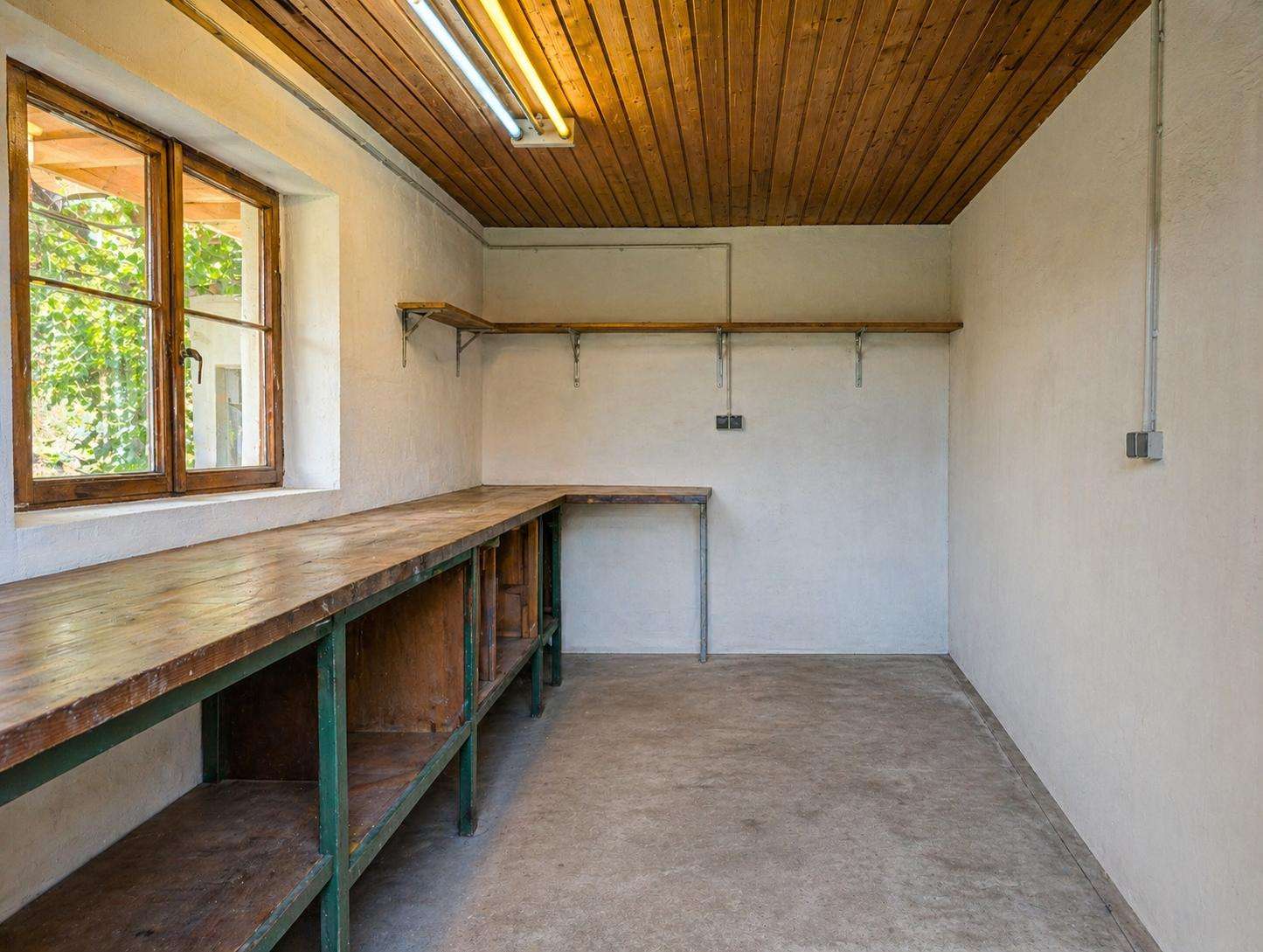 Long wooden workbench runs along the left wall in a simple workshop, with shelves underneath, a window, and an empty, light-filled room.