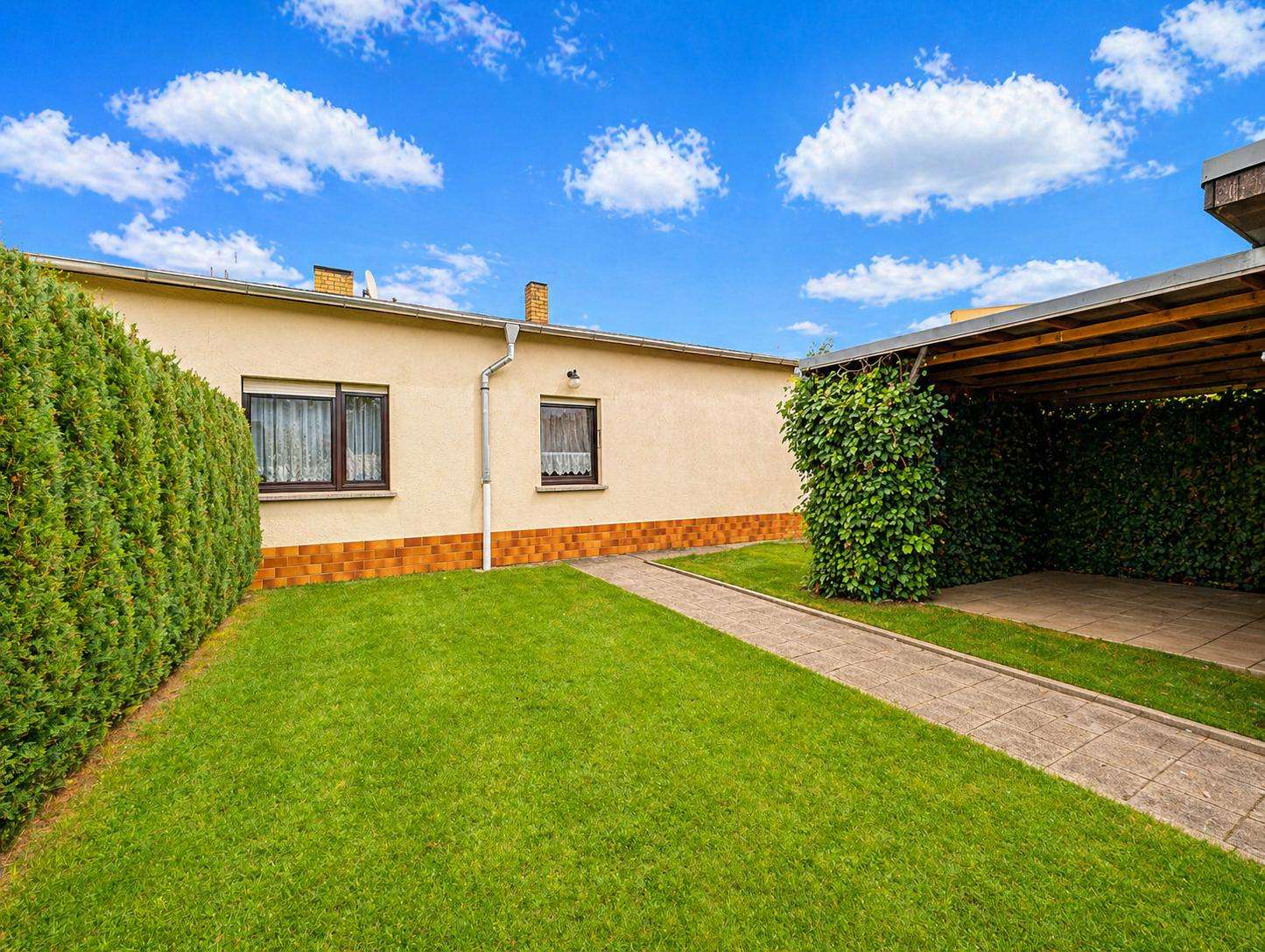 Backyard of a single-story house: green lawn, tall hedges along the left, beige walls with brick base, two brown-framed windows, and a paved path to the side door under a blue sky.