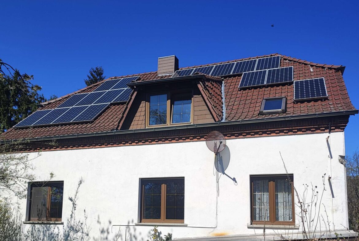 House with multiple solar panels on a red-tiled roof under a clear blue sky, satellite dishes on the wall.