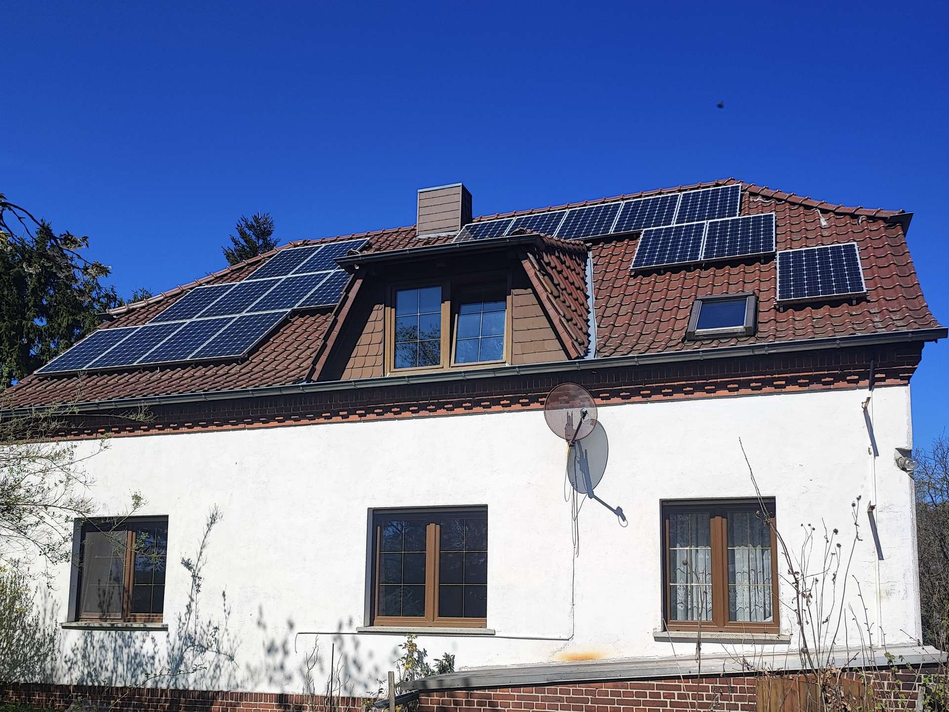 House with multiple solar panels on a red-tiled roof under a clear blue sky, satellite dishes on the wall.