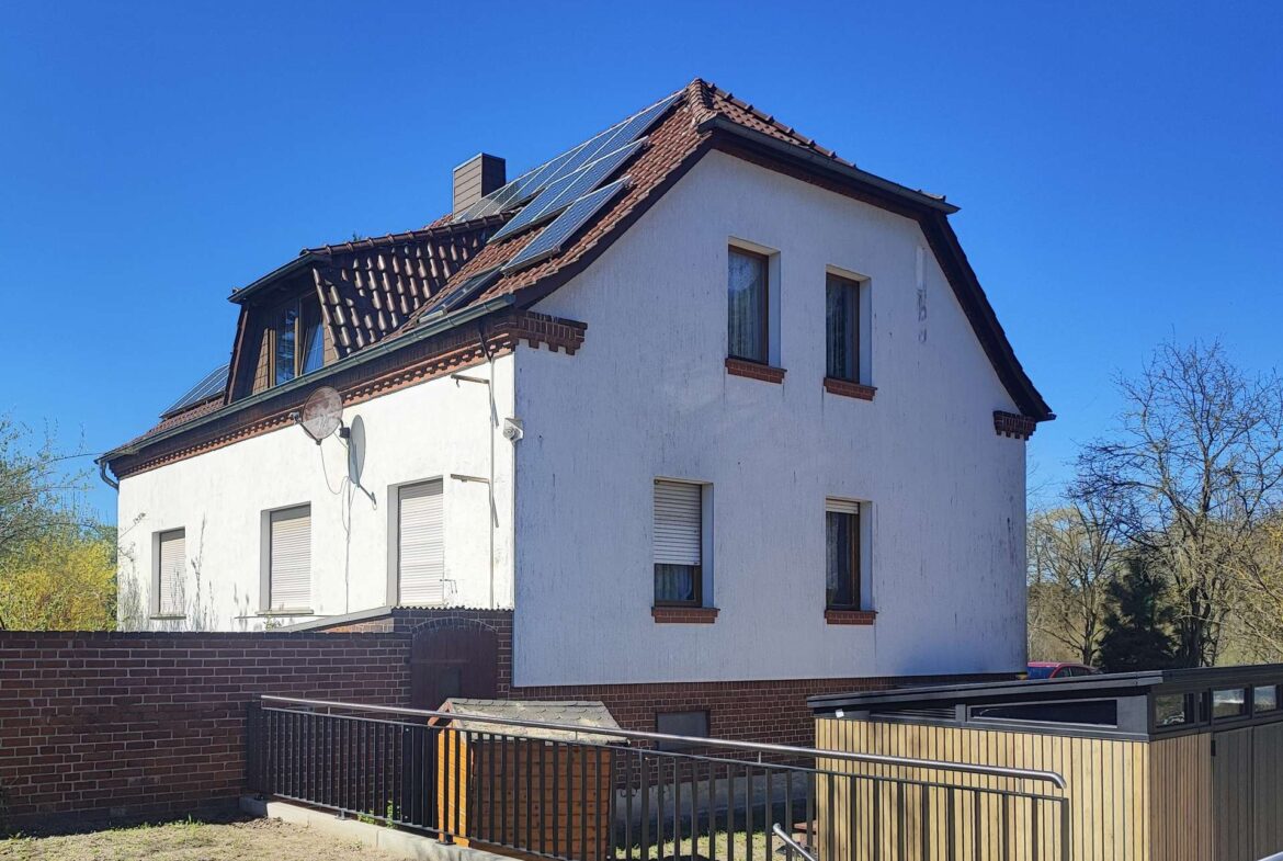 White multi-story house with red-tiled roof and solar panels along the upper slope under a clear blue sky, brick accents and a satellite dish on the wall right side
