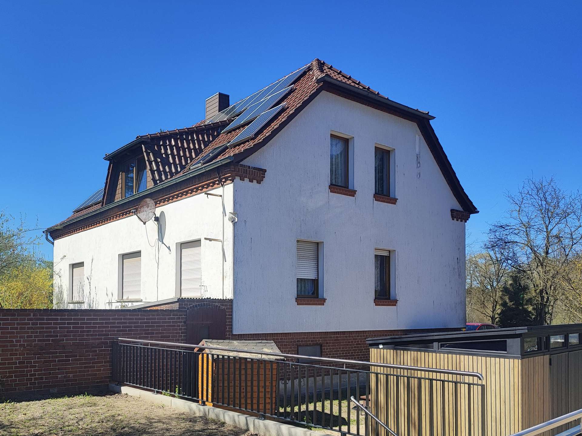 White multi-story house with red-tiled roof and solar panels along the upper slope under a clear blue sky, brick accents and a satellite dish on the wall right side