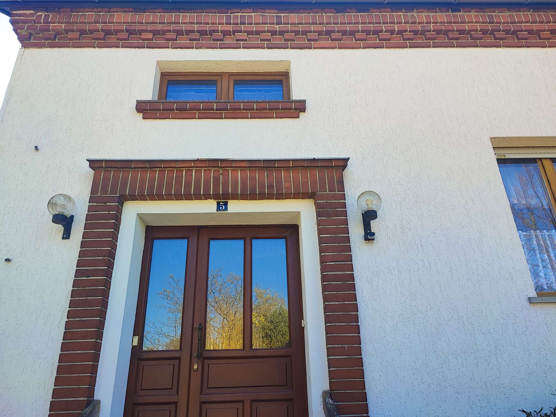 Front door with brown wood panels framed by red brick and white stucco, number 5, with two wall lamps.