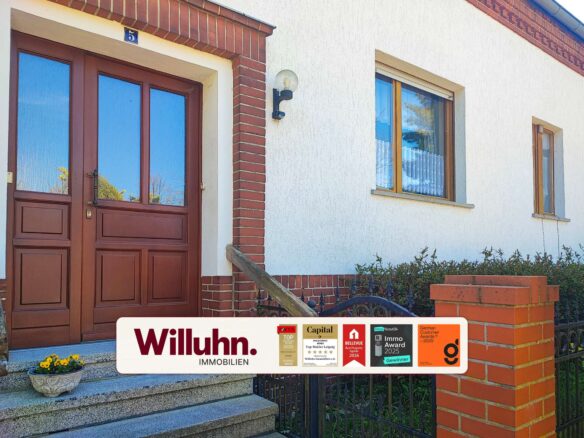 Residential building facade with red brick trim, brown wooden double doors, and a large white real estate banner reading 'Willuhn Immobilien' across the lower portion with award stickers.