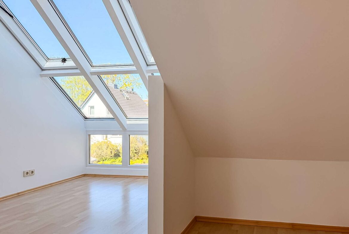 Bright attic room with large angled skylight windows, white walls, and light wood flooring.