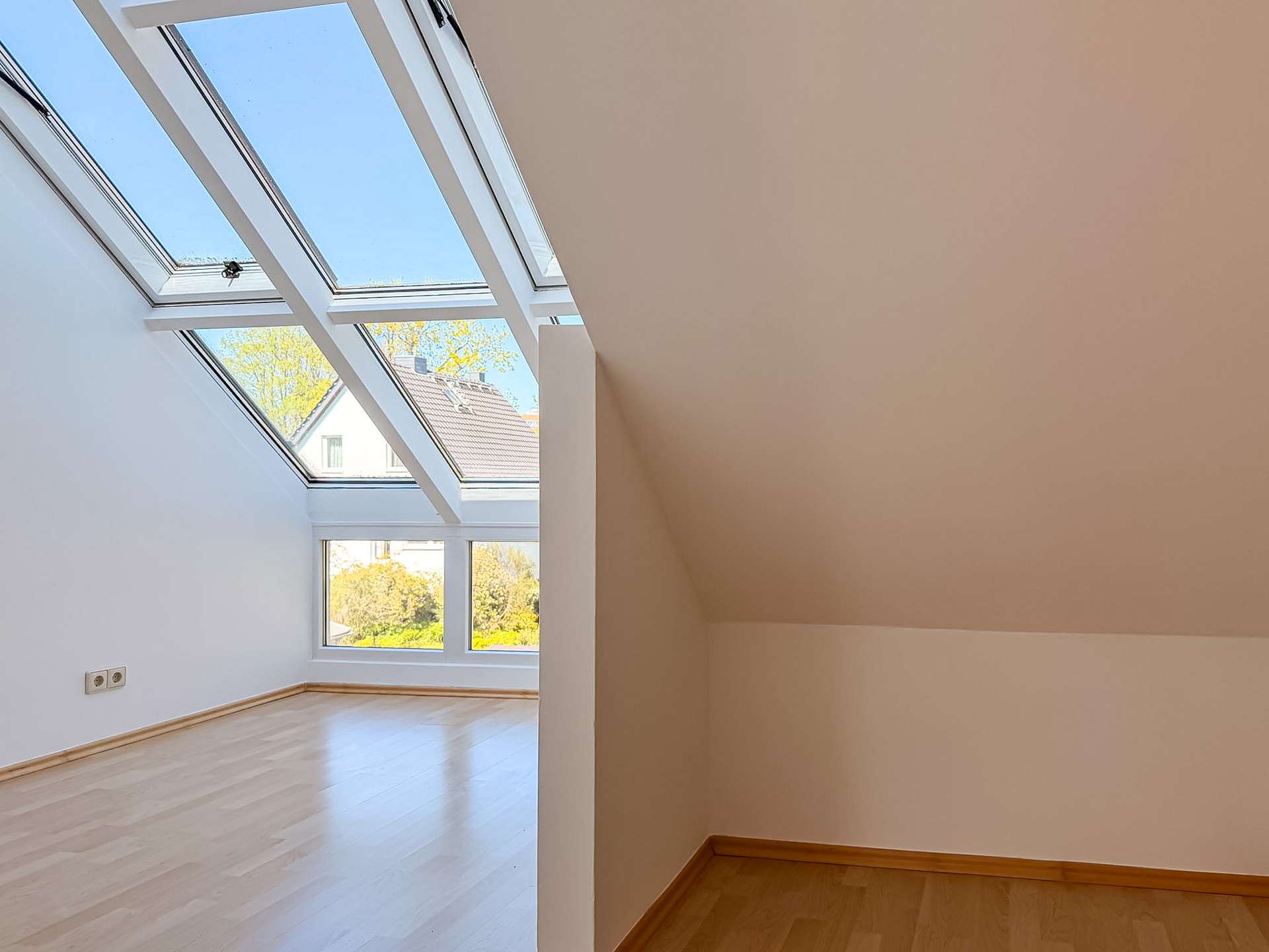 Bright attic room with large angled skylight windows, white walls, and light wood flooring.