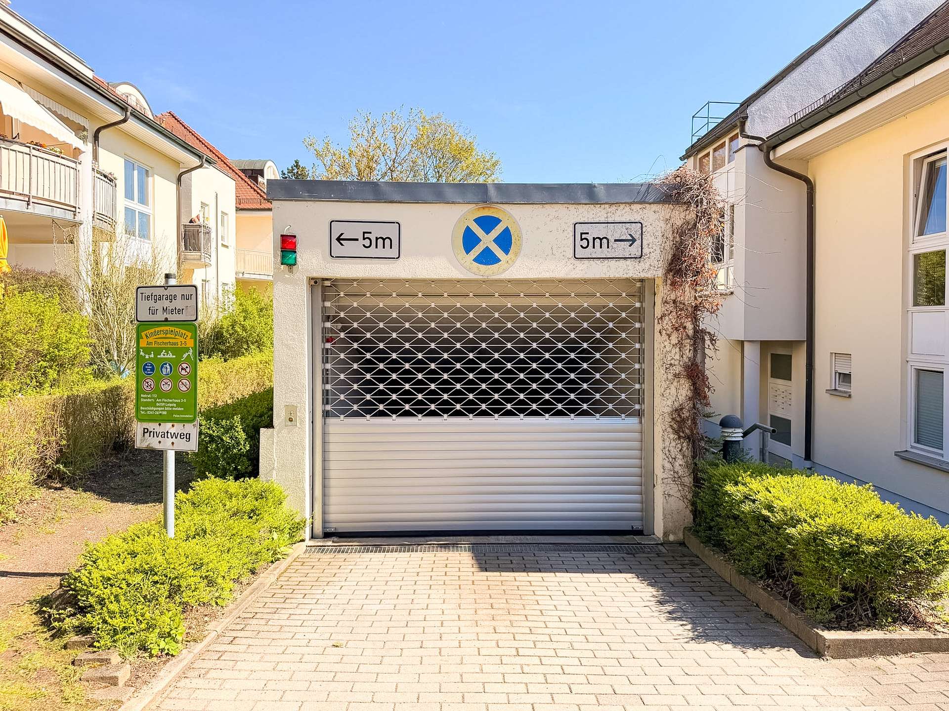 Garage entrance with a metal roll-up door and a no-parking sign above, flanked by residential buildings under a clear blue sky.