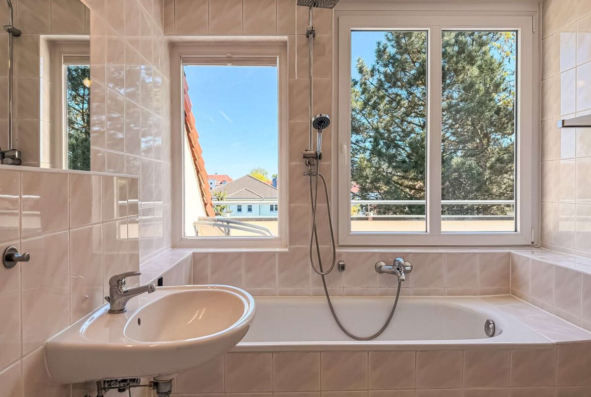 White tiled bathroom with a bathtub, wall-mounted faucet, and a window showing trees outside; handheld shower on the wall and a sink to the left.