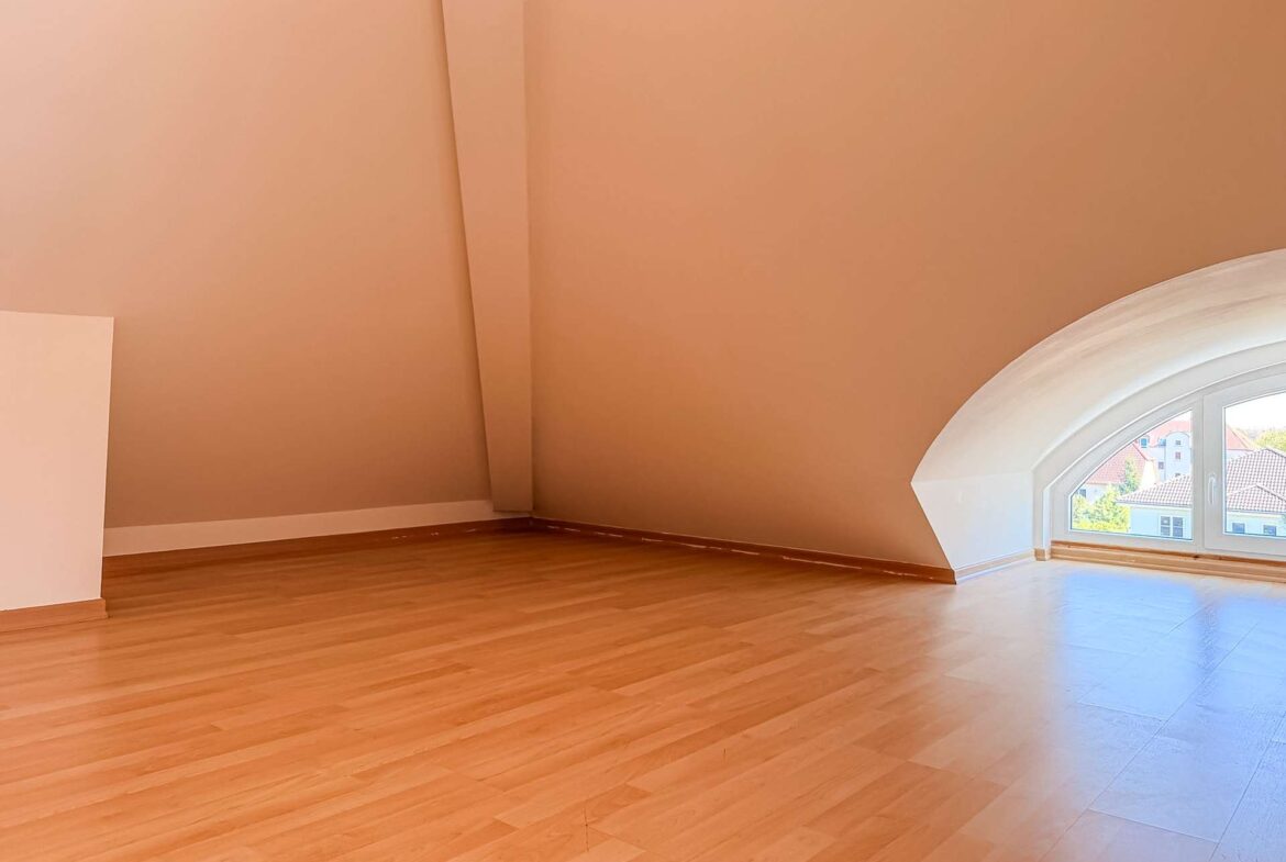 Empty attic room with warm peach walls, light wood floor, and a large arched window letting in sunlight.