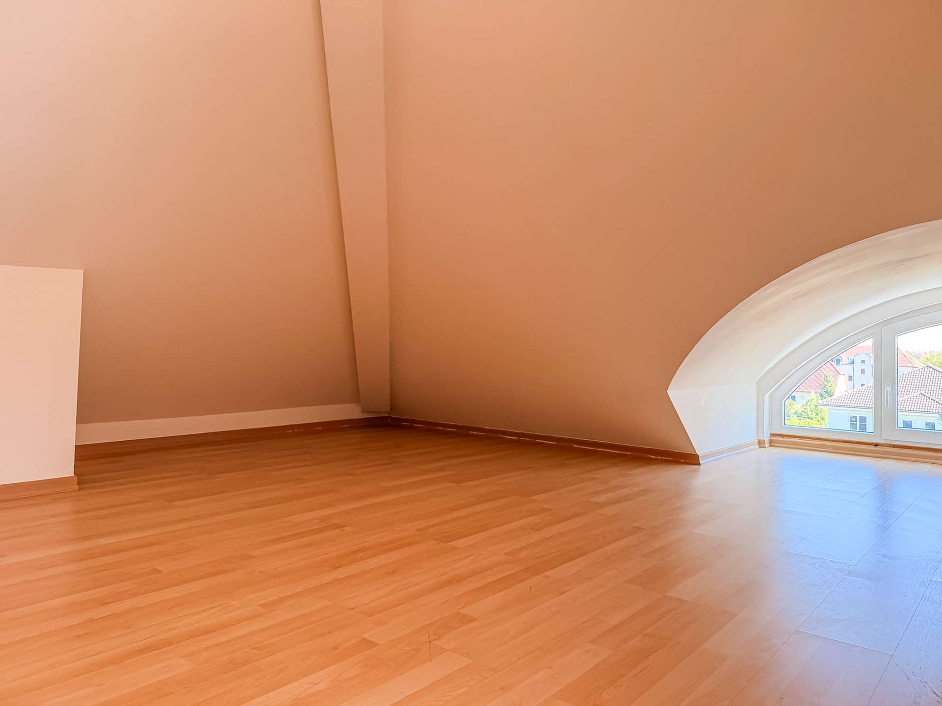 Empty attic room with warm peach walls, light wood floor, and a large arched window letting in sunlight.