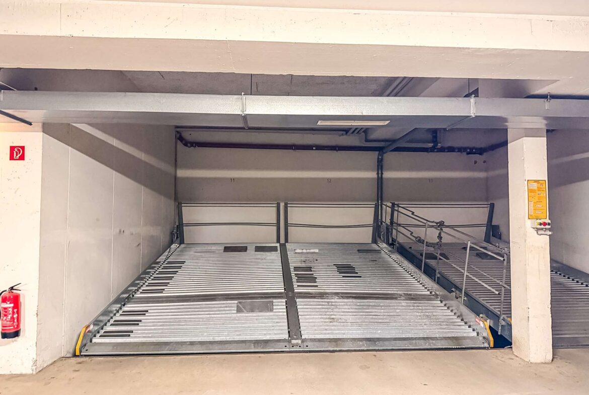 Underside view of a parking garage car stacker with a metal slatted ramp, concrete beams, stairs on the right, and a red fire extinguisher on the left wall.