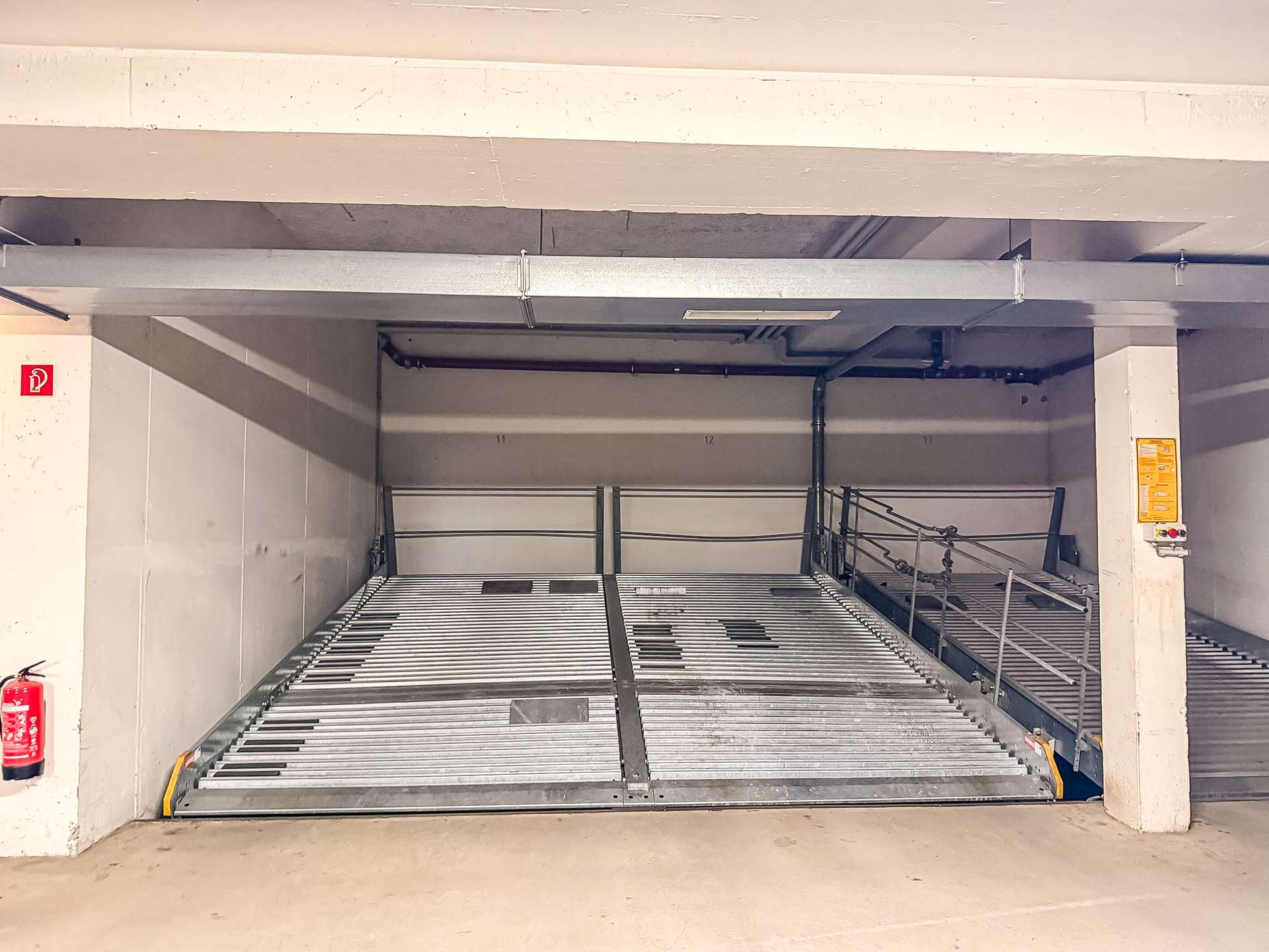 Underside view of a parking garage car stacker with a metal slatted ramp, concrete beams, stairs on the right, and a red fire extinguisher on the left wall.